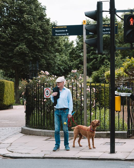 A man standing on a paved sidewalk at a busy street corner holds a leash attached to a brown dog, which is sitting calmly nearby. The man is dressed in a light blue shirt, dark trousers, and a beige f