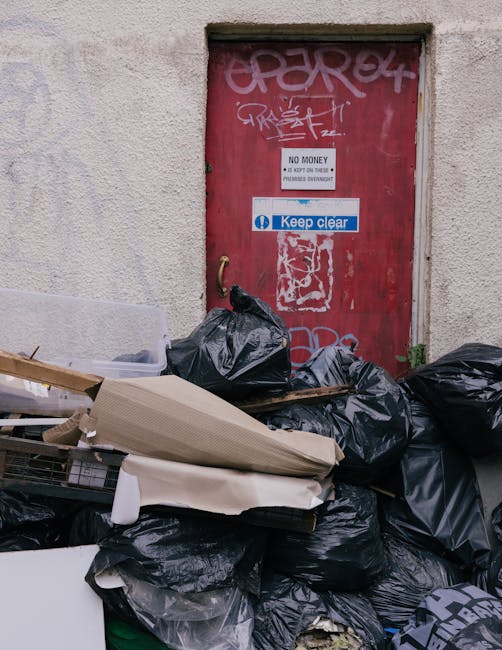 An outdoor scene showing a pile of mixed waste and rubbish bags on a gravel surface beside a low stone wall. The waste includes black plastic bags, some torn open revealing paper and plastic debris, a